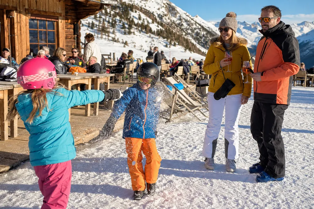Kinder mit Skihelm spielen im Schnee vor einem Bergrestaurant